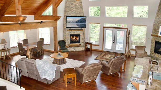 A spacious living room with a stone fireplace, wooden ceiling beams, and large windows offering a view of the outdoors. - Beautiful home on the 10th green on Priest Lake Golf Course (Priest Lake)