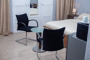 A hotel room with a bed, a glass table, two black chairs, a small desk, and a nightstand with a lamp and a cup. - Studio cosy et confortable (Lomé)
