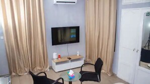 Une chambre d’hôtel dotée d’un téléviseur à écran plat fixé au mur, d’une petite table ronde avec des chaises, d’une table de chevet avec une bouteille et une boîte de mouchoirs, et de rideaux beiges.