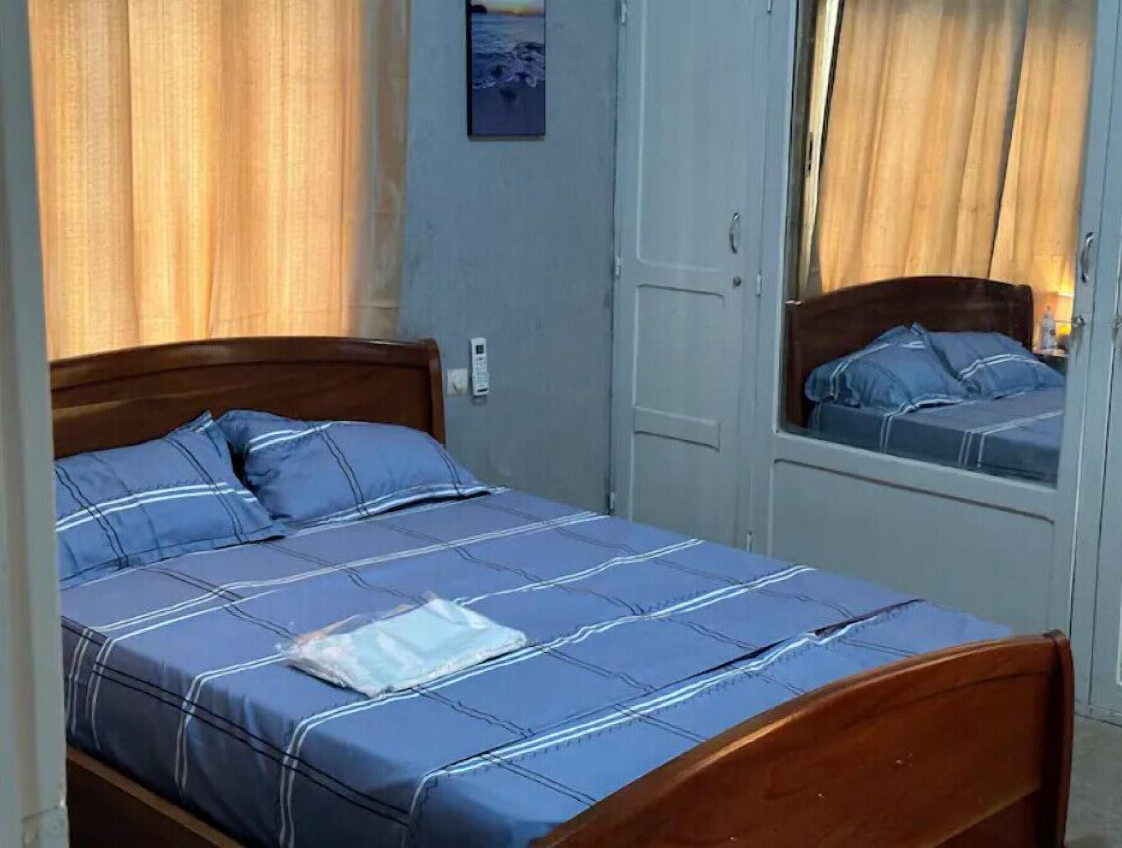 Un lit simple avec une tête de lit en bois, une couvre-lit à rayures bleues et blanches et une petite fenêtre avec des rideaux.