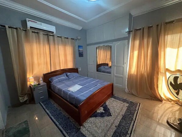 A bedroom with a bed, nightstands, an air conditioning unit, a window with curtains, and a door with a view.