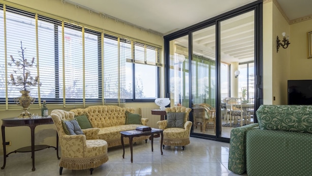 A spacious living room with a sofa, armchairs, a coffee table, and a TV. Large windows with blinds provide natural light. - GuestHost - Grazia Attic Apartment with terrace (Trani)