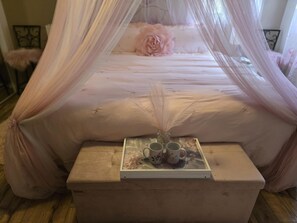 A bedroom with a canopy bed, a bedside table with two cups and a vase, and a fluffy pillow on the bed.