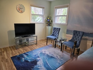 A living room with a flat-screen TV on a glass table, two blue armchairs, a rug with a wave design, and a clock with seashells on the wall.