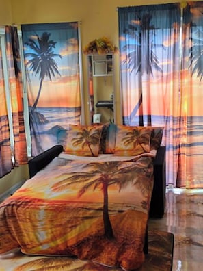 Une chambre Ă coucher avec un lit, des rideaux ornĂ©s dâun motif reprĂ©sentant un coucher de soleil tropical, une table de chevet et un miroir.