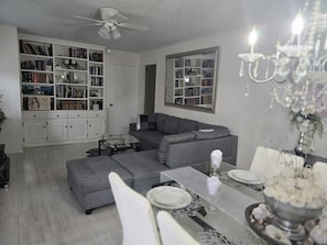 A modern living room with a grey sofa, a glass coffee table, and a dining table set for four. There is a bookshelf filled with books and decorative items, and a chandelier hangs from the ceiling.