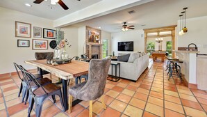 A spacious living area with a wooden dining table, grey upholstered chairs, a grey sofa, a fireplace, and a TV mounted on the wall.