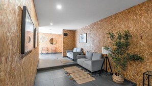 Un interior moderno con un sofá gris, una mesita y una lámpara. Hay dos sillas y una planta. Las paredes están revestidas con un material que imita a la madera.