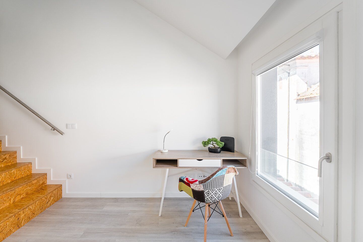 A modern, minimalistic room with a wooden staircase, a desk with a plant, and a chair with a colorful cushion.