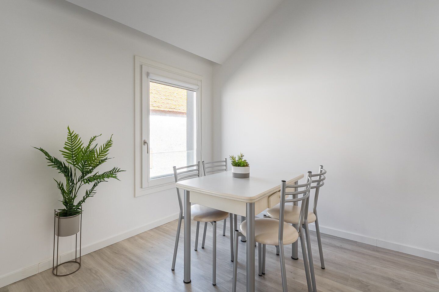 Et spiseområde med et hvidt bord, fire stole, en potteplante og et vindue med skodder.