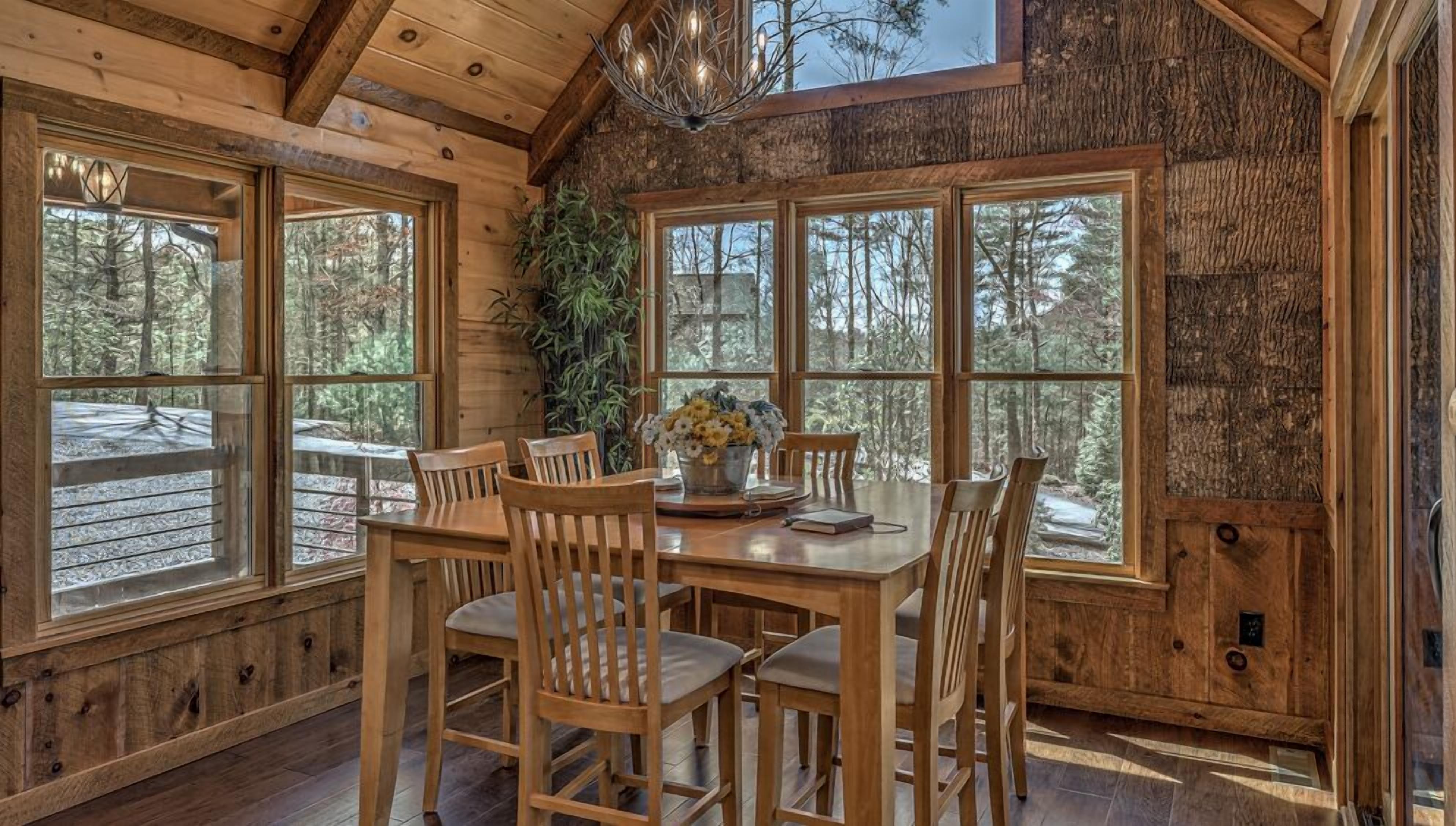 A wooden dining area with a table and chairs, large windows with a forest view, and a chandelier.