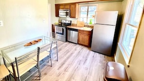 Cocina compacta con gabinetes de madera, refrigerador de acero inoxidable, microondas, horno y lavavajillas. Hay una pequeña mesa de comedor con sillas.
