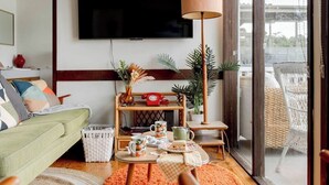 Ein Wohnzimmer mit grünem Sofa, einem Couchtisch mit Teeservice, einem an der Wand montierten Fernseher und einem Korbstuhl am Fenster.