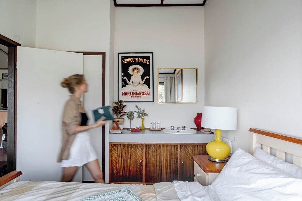 A hotel room with a bed, a bedside table with a lamp, a mirror, a vanity with a sink, and a poster on the wall.