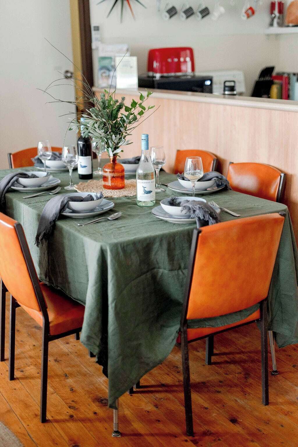 A dining table set with plates, glasses, and a bottle of wine, surrounded by orange chairs.