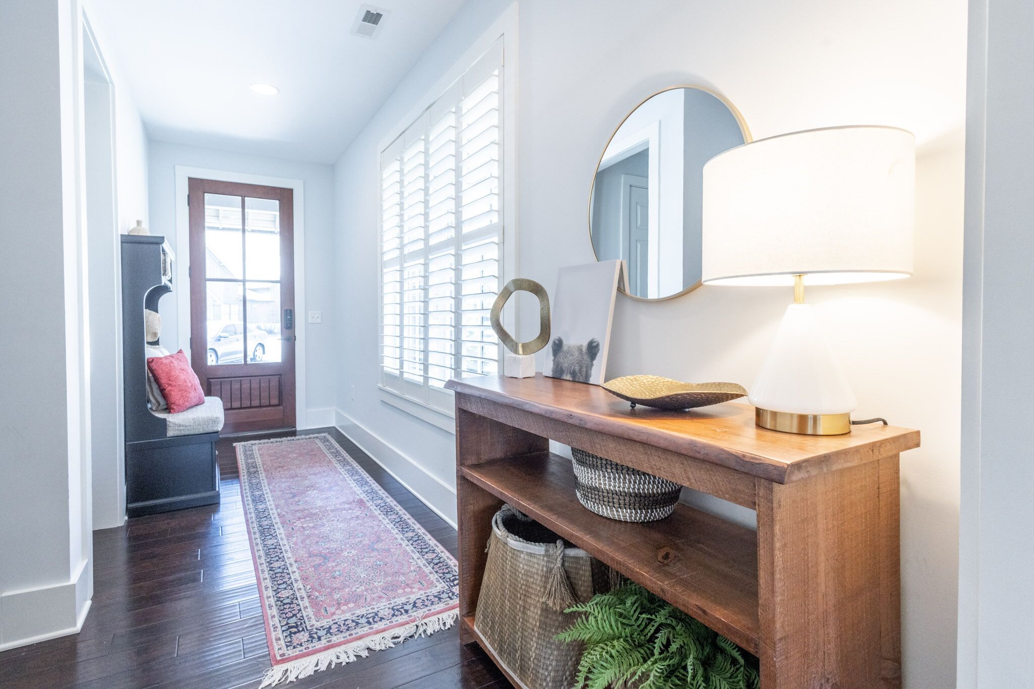 Un couloir avec une console en bois, un miroir et une lampe. On y trouve une porte avec un détail rouge, un banc et un tapis à motifs.