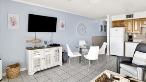A modern living room with a flat-screen TV, a dining area with a round table and white chairs, a kitchen with wooden cabinets, and a ceiling fan. - Beachfront @ Seaside Beach & Racquet 1BR • Sleeps 4 • Amazing Views (Orange Beach)