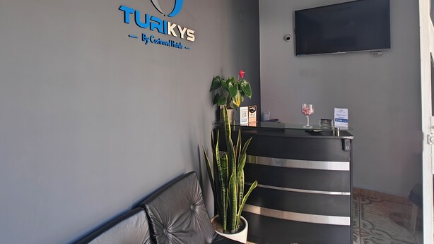 Ein Empfangsschalter mit dem Logo von 'TURKYS By Controversy Hotels', ein an der Wand montierter Flachbildfernseher und ein Blumentopf neben dem Schalter.