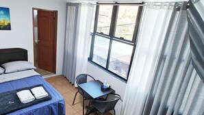 Ein Hotelzimmer mit einem Bett, einem Tisch und einem Stuhl. Es gibt ein Fenster mit Vorhängen, eine Tür und ein Bild an der Wand.