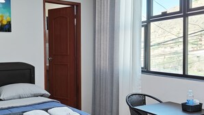 Ein Schlafzimmer mit einem Bett, einem Blumenbild, einer Holztür, einem Fenster mit Blick auf Gebäude und einem Tisch mit einer Flasche.
