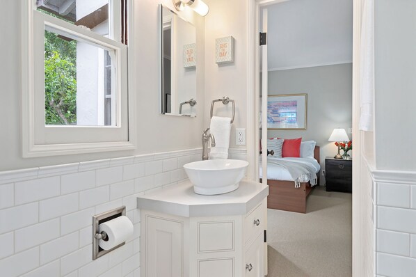 Un baño con un lavamanos blanco, un espejo y una ventana. Hay una puerta que da a un dormitorio con una cama y una mesita de noche con una lámpara.