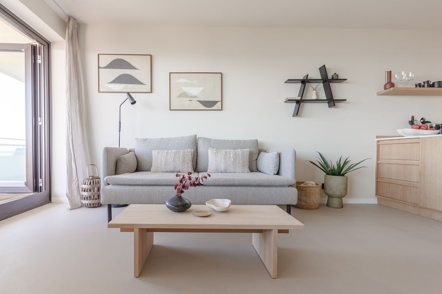 A modern living room with a grey sofa, a wooden coffee table, and a minimalist decor including abstract wall art and a ladder shelf.