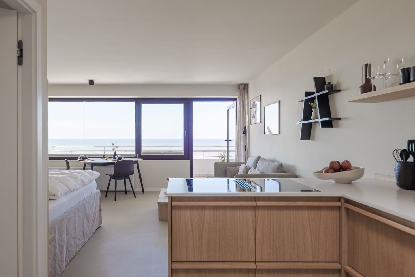 Une chambre d’hôtel moderne dotée d’une grande fenêtre donnant sur la mer, d’une kitchenette avec des placards en bois, d’un coin repas avec une table et des chaises, et d’un coin salon confortable avec un canapé.