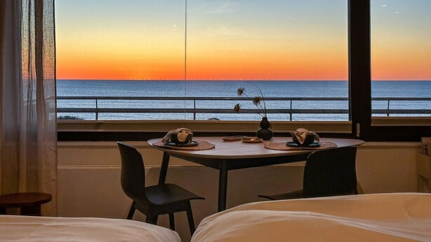 A room with a view of the sunset over the ocean, a dining table with plates and a vase, and two beds. - Appartement Ocean Suite - Ocean Suite (Wenningstedt-Braderup (Sylt))