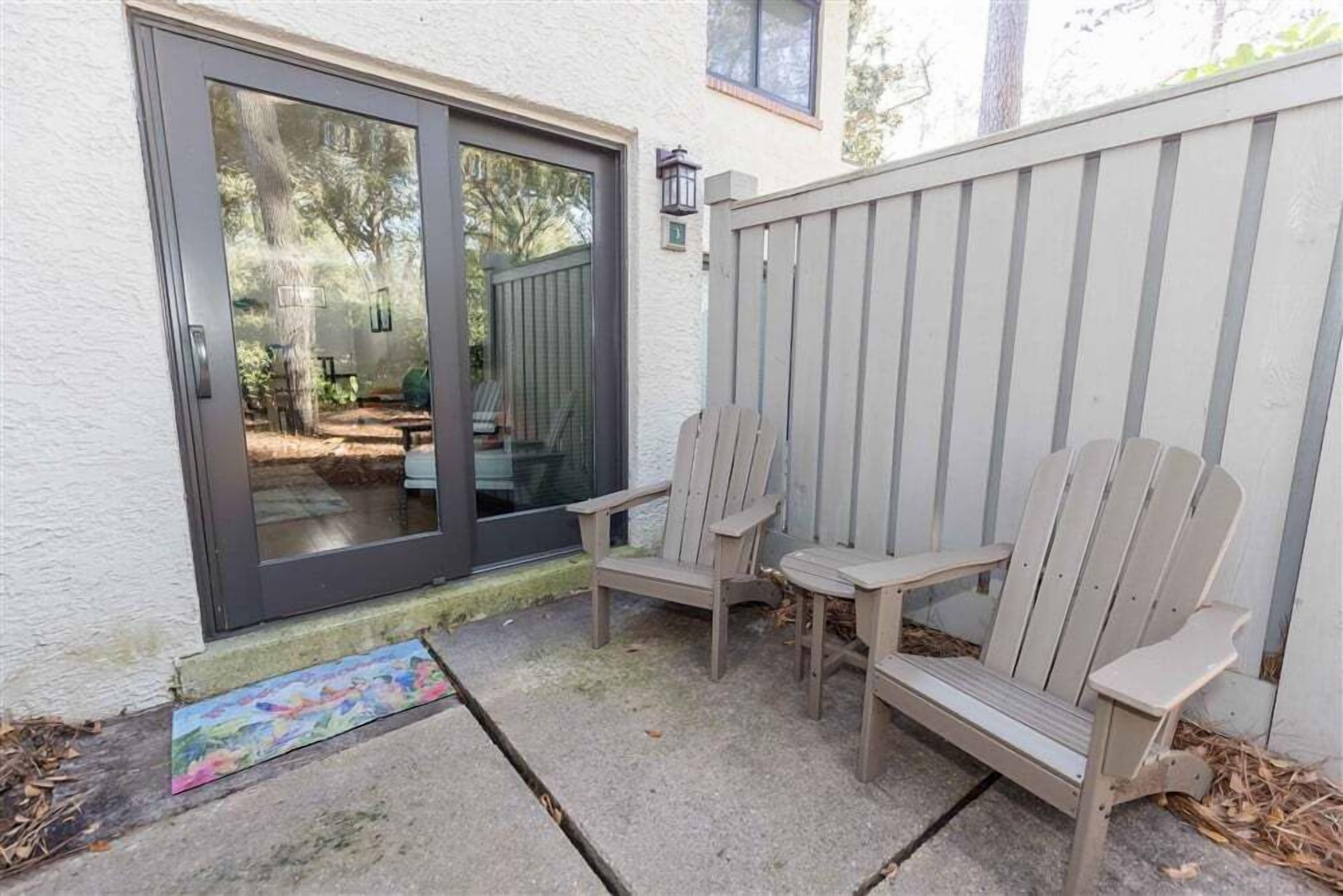 En patio med to Adirondack-stole og et lille bord, en skydedør og et træhegn.
