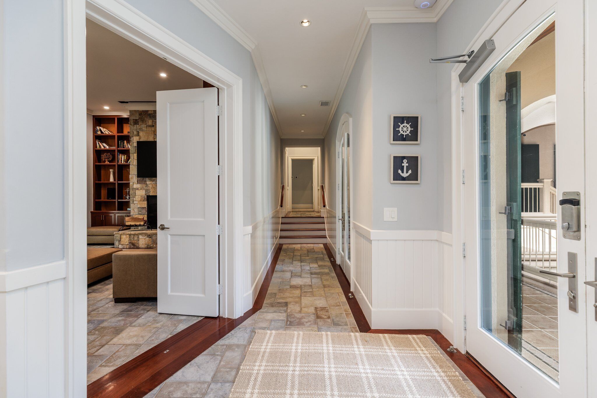 A hallway with a stone floor, a patterned rug, a staircase, and a bookshelf with books and decorative items.