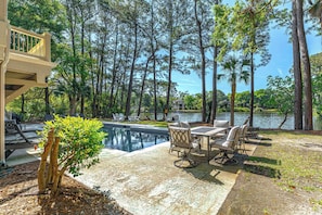 A pool area with a view of a lake, surrounded by tall trees and a patio with outdoor furniture.