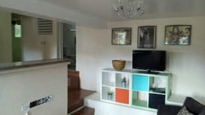 A living room with a television on a white cabinet, a chandelier, and framed pictures on the wall. - TonLin Pewee Drive 1 Bedroom (Gros Islet)