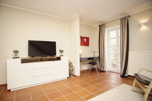 A living room with a TV on a white cabinet, a desk with a chair, and a window with curtains.