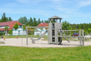 A playground with a wooden climbing structure and slides, surrounded by a grassy area and a building with a red roof in the background. - Die Ferienwohnung Faro im Feriendorf Bakenberg - Feriendorf Bakenberg (Dranske)