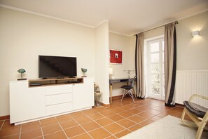 Een woonkamer met een televisie op een witte kast, een bureau met een stoel, en een raam met gordijnen.