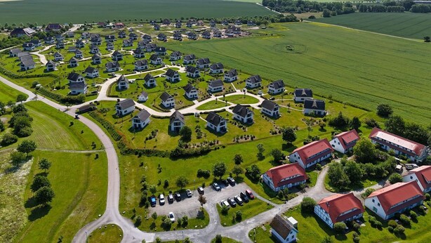 Vogelperspektive eines Wohngebiets mit in Kreismuster angeordneten Häusern, umgeben von grünen Feldern und einigen größeren Gebäuden mit roten Dächern.
