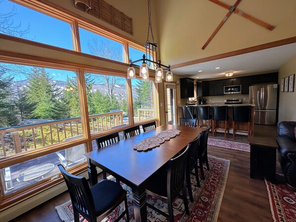A dining area with a large table and chairs, a kitchen in the background, and a view of trees and mountains through large windows.