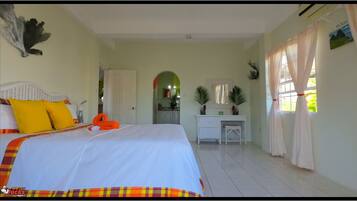 A bedroom with a bed, a desk, and a view of the kitchen.