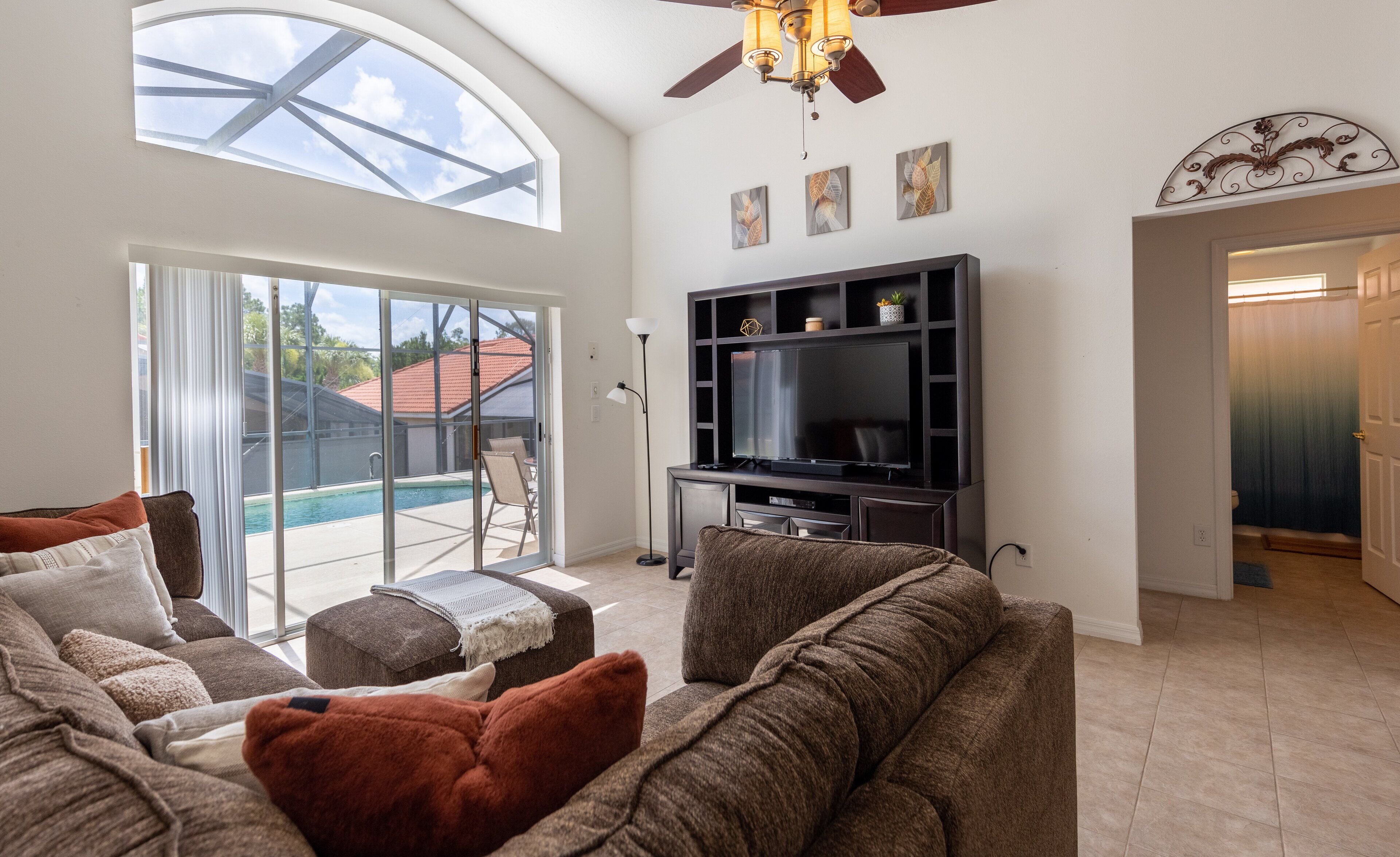 A living room with a large TV, comfortable seating, a ceiling fan, and a view of a swimming pool.