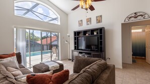A living room with a large TV, comfortable seating, a ceiling fan, and a view of a swimming pool. - Aviana resort near Disney, private pool, WiFi, washer, dryer, gameroom (Davenport)