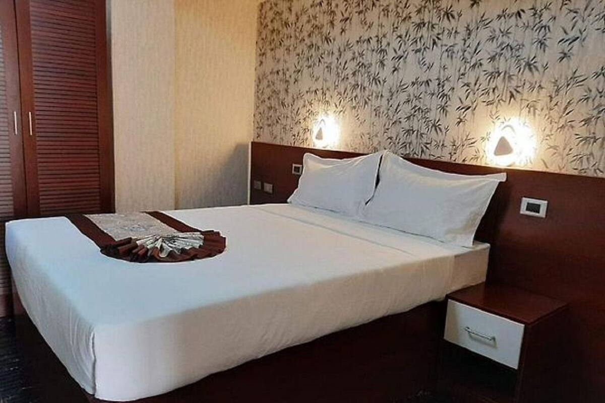 A neatly made bed with a white bedspread and two pillows, a wooden nightstand with a drawer, and a wall with a floral wallpaper pattern.