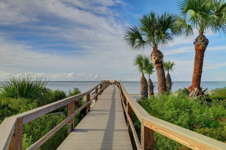 Wooden boardwalk leading to the beach at Skidaway Island State Park near Savannah, Georgia, surrounded by lush greenery and palm trees under a bright blue sky