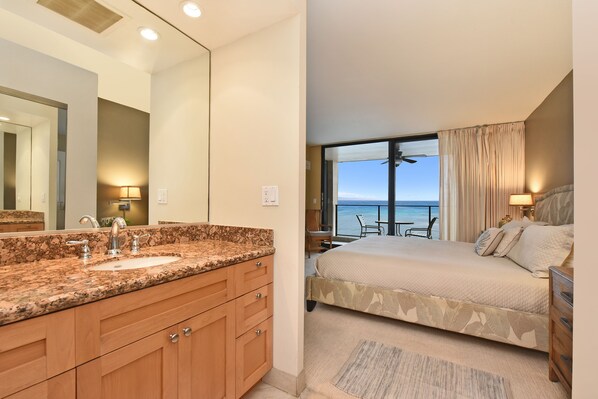 A hotel room with a large bed, a bathroom with a granite countertop sink, and a balcony with a view of the ocean.