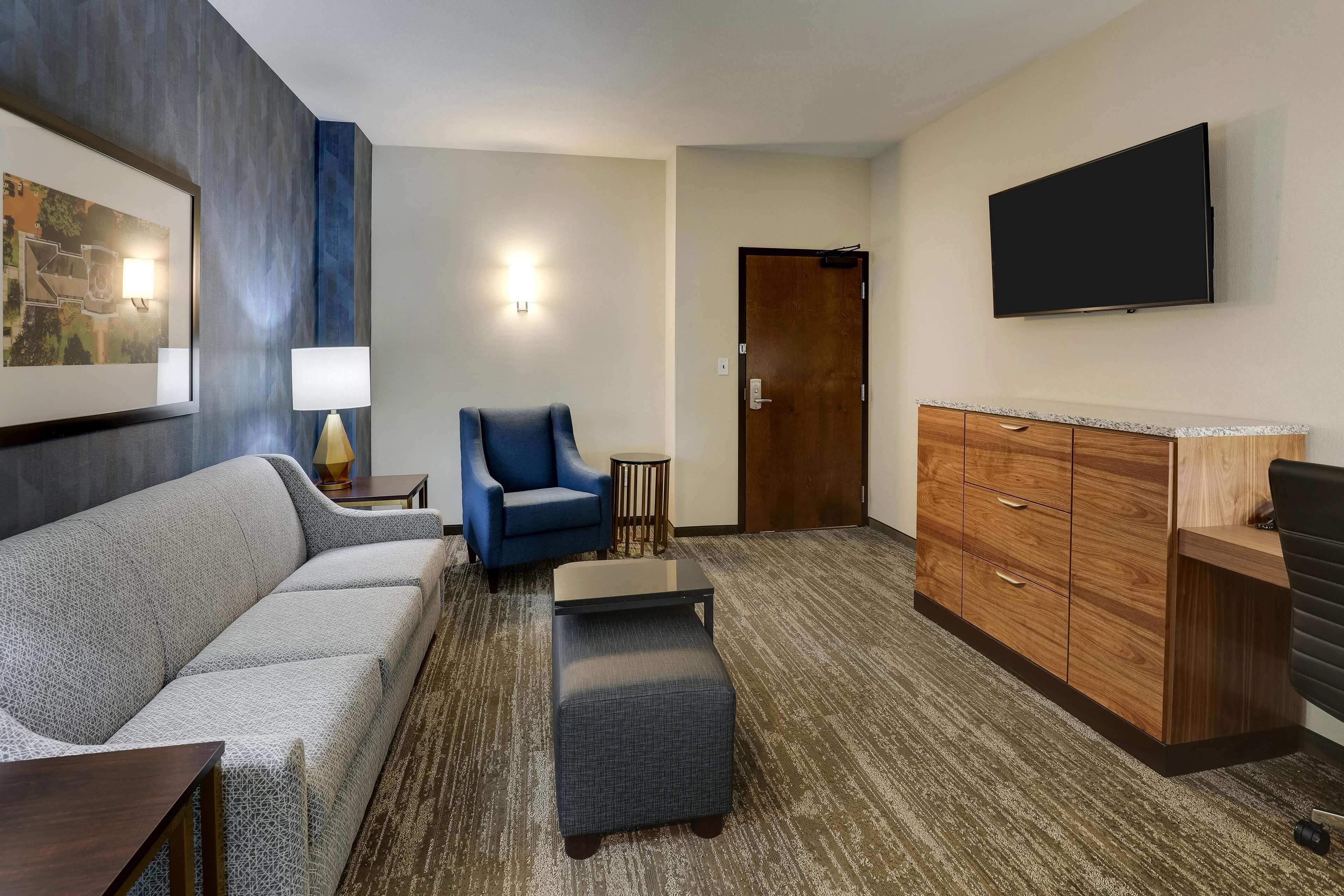 Una habitación de hotel moderna con un sofá gris, un sillón azul, una mesita pequeña, un aparador de madera y un televisor de pantalla plana montado en la pared.
