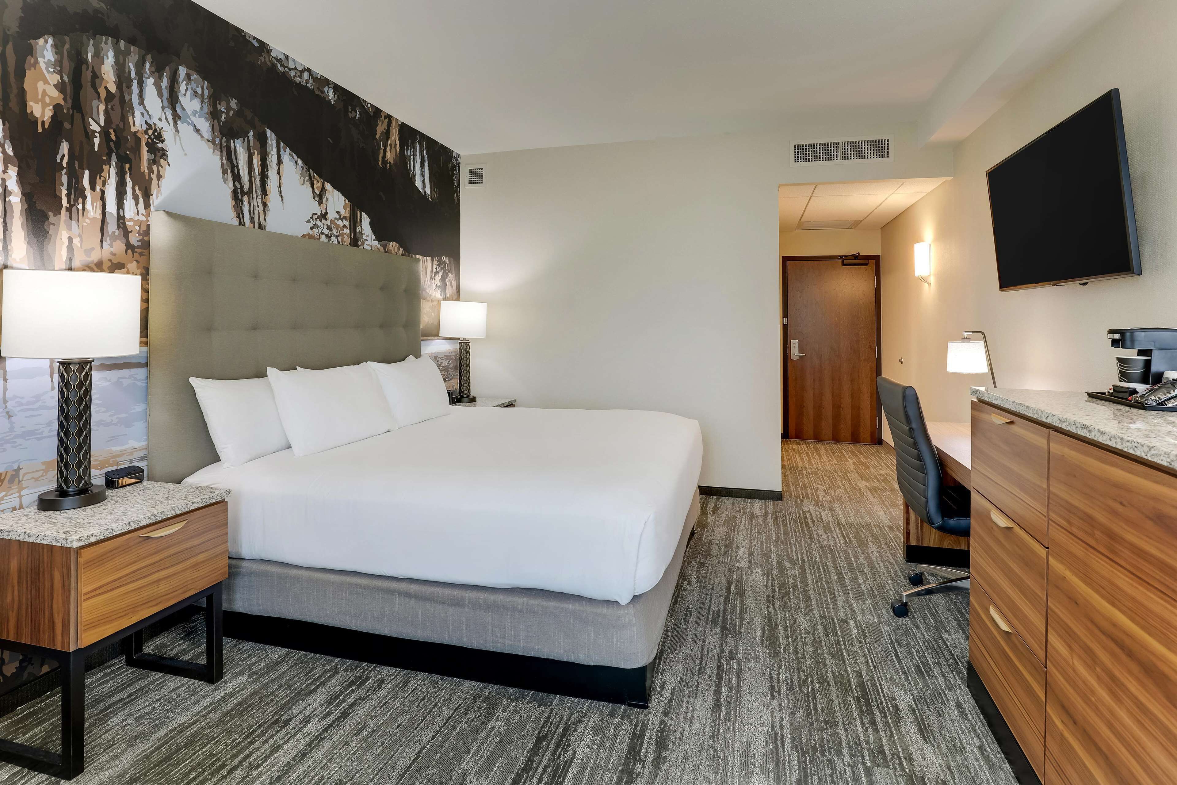 Une chambre d’hôtel avec un grand lit, deux tables de chevet avec des lampes, un bureau avec une chaise, un téléviseur à écran plat et un grand tableau accroché au mur.