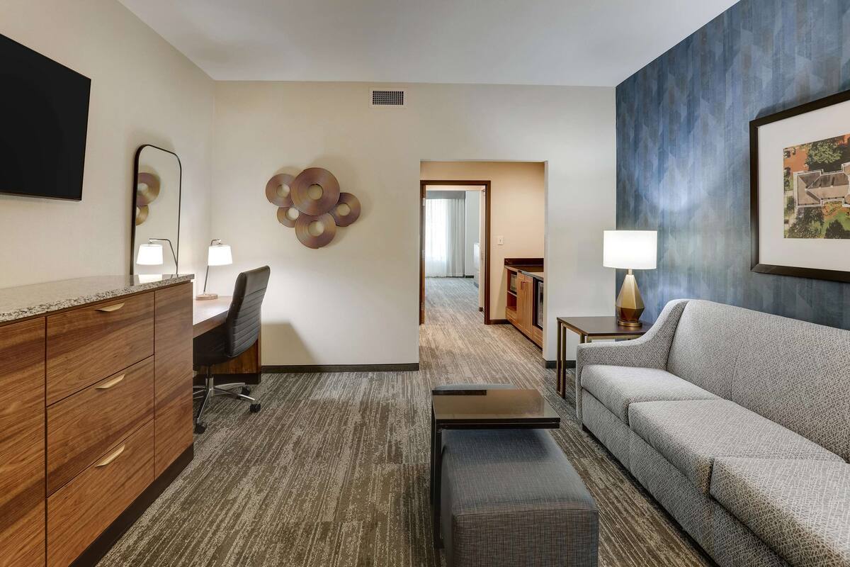 A hotel room with a wooden dresser, a gray sofa, a black office chair, a small table, and a framed picture on the wall.