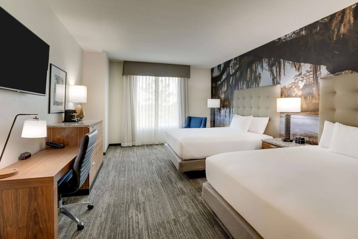 A hotel room with two beds, a desk, a chair, a TV, and a large window with curtains.