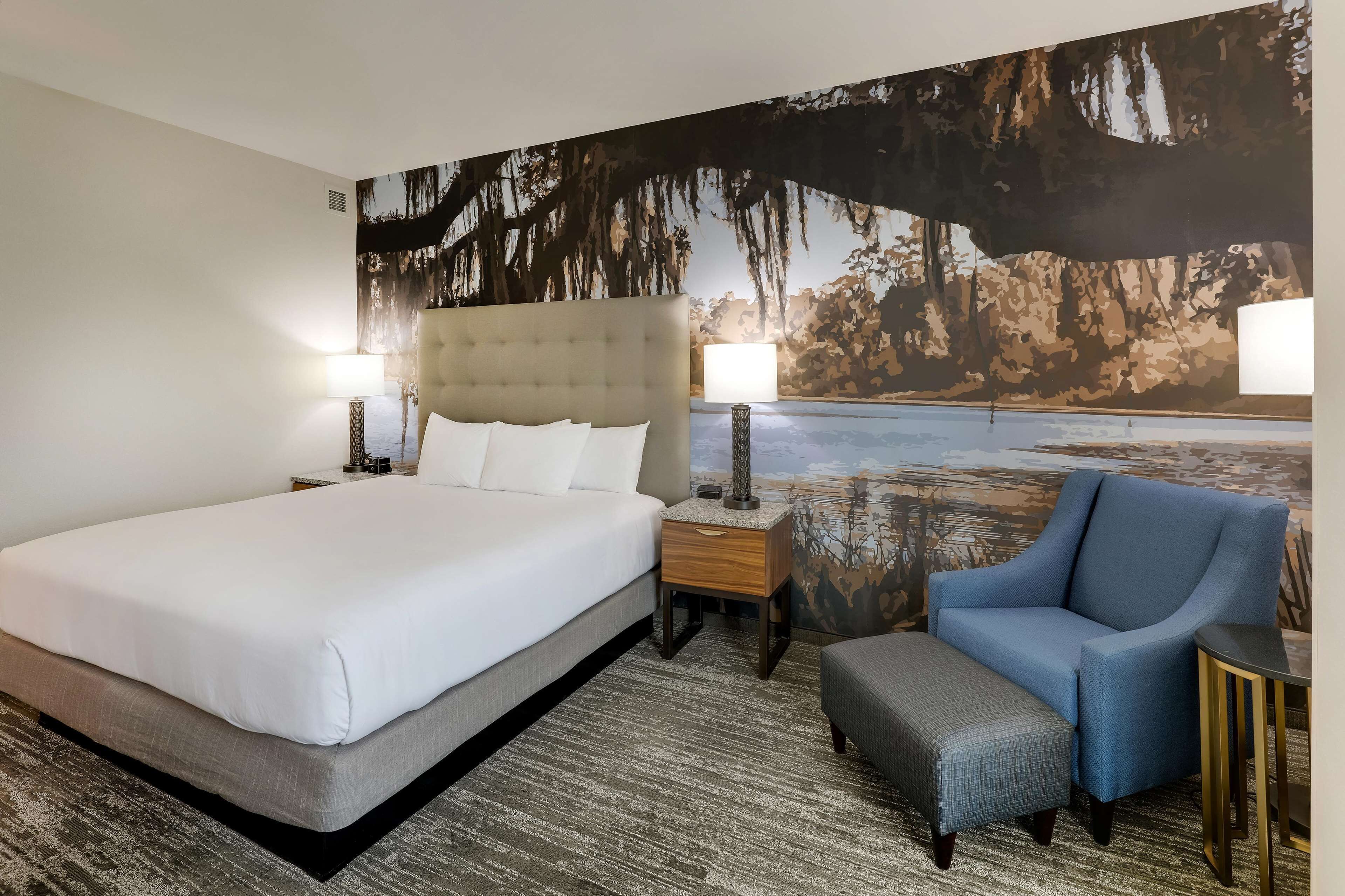 Habitación de hotel con una cama grande, un sillón azul, una mesita y una mesita de noche con lámparas.