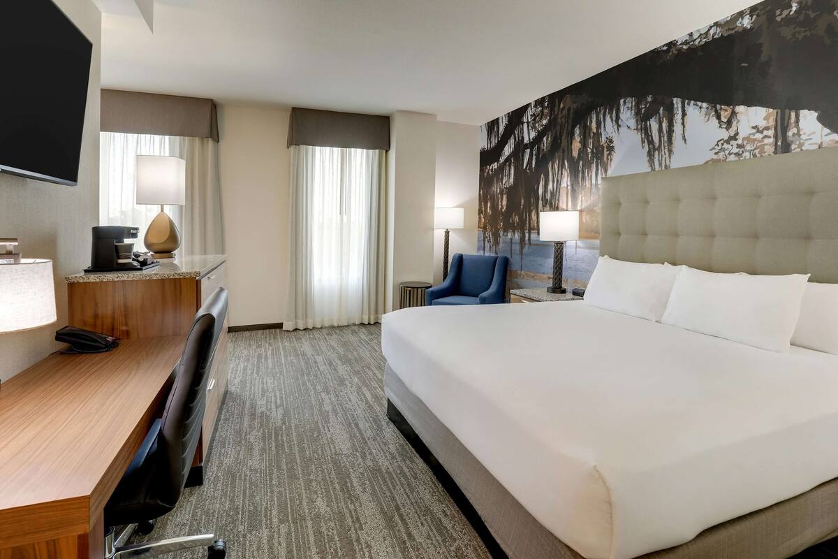 A hotel room with a large bed, a desk with a chair, a television, and a painting on the wall.