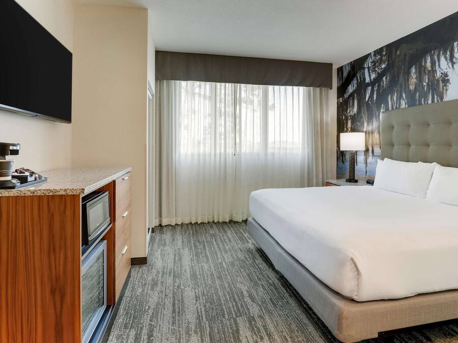 Una habitación de hotel con cama, televisión, cafetera y un ventanal con cortinas.
